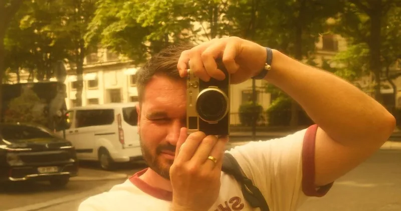 Sam Buckingham raising a compact camera to one eye on a tree-lined city street, in warm golden light